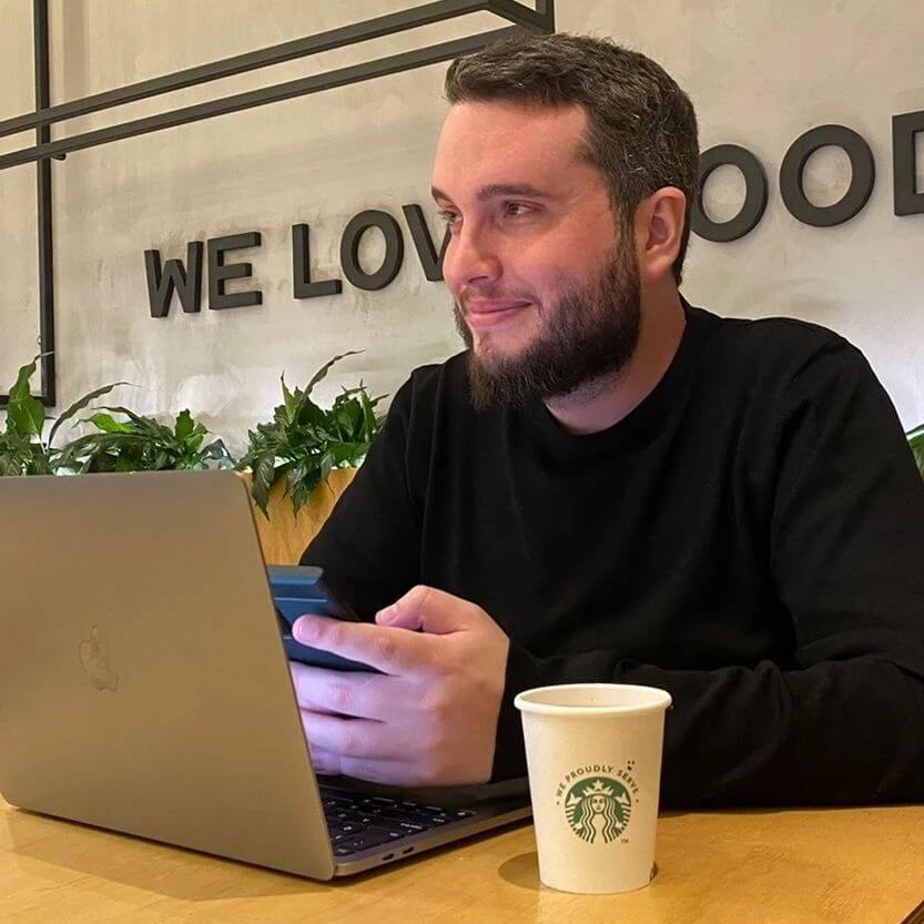 Felipe Goulart sorrindo em um café, trabalhando com notebook e celular